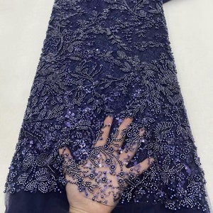 May include: A close-up of a navy blue beaded lace fabric with a floral pattern. The fabric is embellished with small, round beads and sequins.