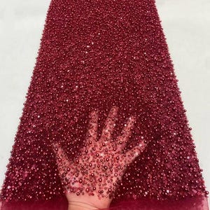 Può includere: Tessuto bordeaux con perline. Il tessuto è densamente ricoperto di piccole perline rotonde di varie dimensioni. Le perline creano una superficie strutturata. Il tessuto è probabilmente utilizzato per abbigliamento o scopi decorativi.