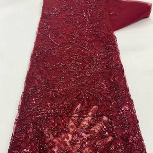 Könnte beinhalten: Ein tiefburgunderroter Spitzenstoff mit aufwendigen Blumenstickereien, zarten Perlenarbeiten und über das Netzmaterial verteilten Pailletten. Das Textil zeigt ein dichtes, verziertes Muster aus Ranken und Blüten in einem passenden Dunkelrot.