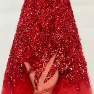 Puede incluir: Una tela roja vibrante con intrincados adornos de cuentas y lentejuelas. El diseño presenta patrones arremolinados y una superficie texturizada, creando una apariencia lujosa y llamativa. Este textil es adecuado para crear ropa elegante.