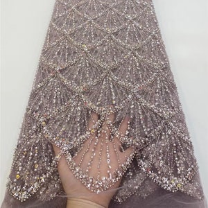 Puede incluir: Primer plano de una tela de color marrón claro con cuentas y un patrón de conchas. La tela está adornada con lentejuelas y cuentas, creando un efecto brillante.