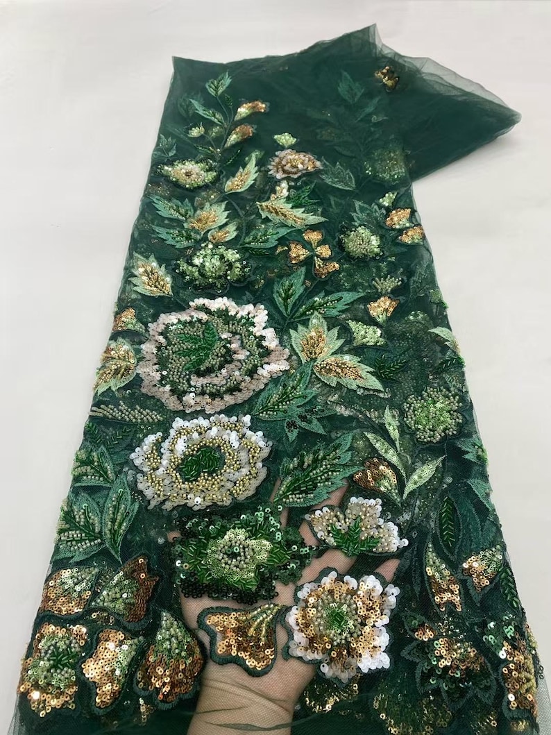 May include: Dark green fabric with intricate floral embroidery. The design features large, detailed flowers in white, gold, and green, with accents of sequins and beads. The fabric has a sheer, net-like appearance.