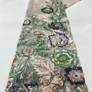 May include: Delicate floral lace fabric featuring intricate embroidery and sequin embellishments. The design includes detailed flowers in shades of green, gold, blue, and brown, with white and green leaves. The sheer fabric is a light beige color.