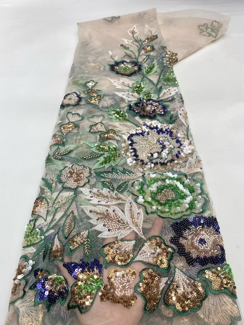 May include: Delicate floral lace fabric featuring intricate embroidery and sequin embellishments. The design includes detailed flowers in shades of green, gold, blue, and brown, with white and green leaves. The sheer fabric is a light beige color.