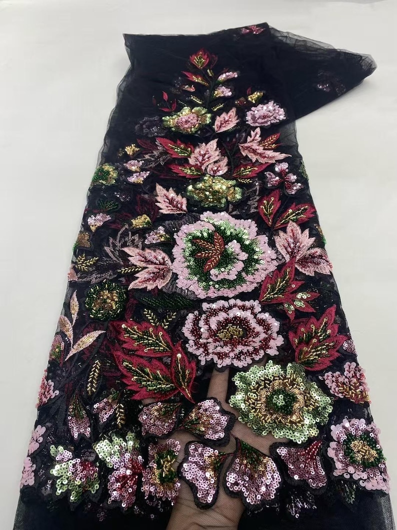 May include: Black fabric with an embroidered floral design. The design features large flowers in shades of pink, green, and red, with gold and green sequins. The fabric is likely intended for use in apparel or decorative projects.