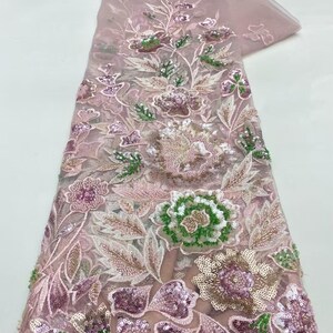 May include: A delicate, sheer fabric in a soft pink hue, adorned with intricate floral embroidery. The design features detailed flowers in shades of pink, green, and gold, embellished with sequins. The fabric is ideal for creating elegant apparel or decorative items.