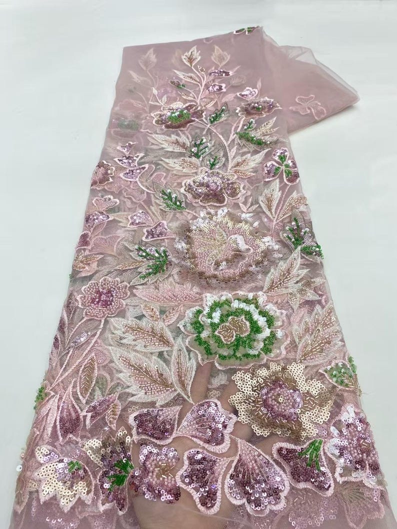May include: A delicate, sheer fabric in a soft pink hue, adorned with intricate floral embroidery. The design features detailed flowers in shades of pink, green, and gold, embellished with sequins. The fabric is ideal for creating elegant apparel or decorative items.