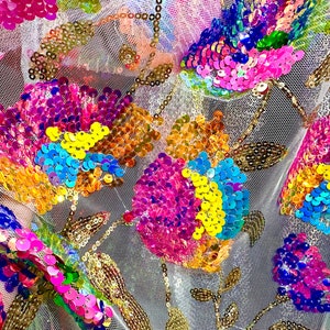 May include: A close-up of a white mesh fabric with colorful sequins in the shape of flowers. The sequins are iridescent and have a variety of colors, including pink, blue, yellow, and green.
