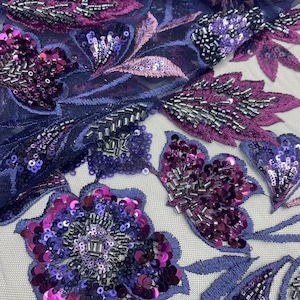 May include: Close-up of a fabric with a floral design. The fabric features embroidered flowers and leaves in shades of purple, magenta, and silver. The flowers are embellished with sequins and beads, creating a textured and detailed appearance.