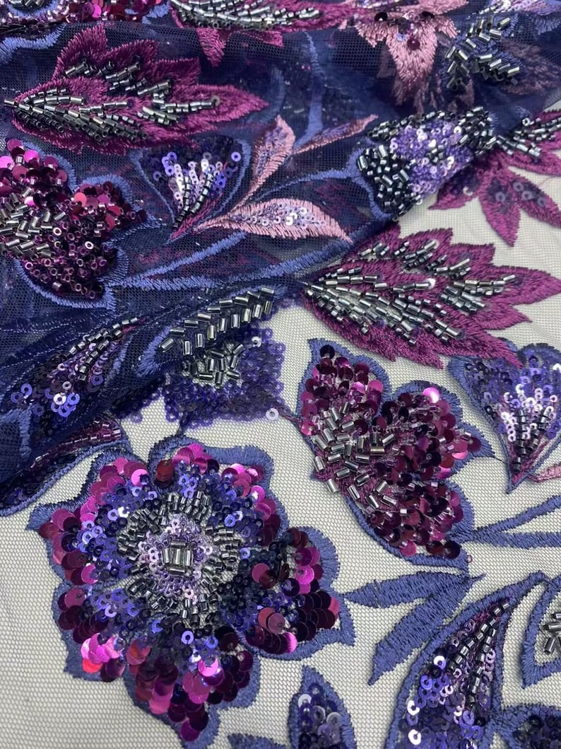 May include: Close-up of a fabric with a floral design. The fabric features embroidered flowers and leaves in shades of purple, magenta, and silver. The flowers are embellished with sequins and beads, creating a textured and detailed appearance.