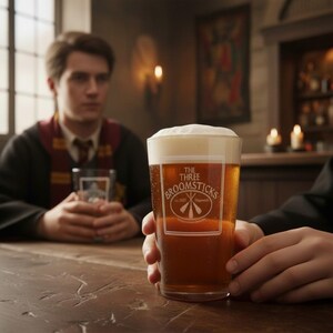 Puede incluir: Un vaso de cerveza rubia clara con una espesa espuma blanca. El vaso lleva la inscripción "THE THREE BROOMSTICKS". La escena tiene lugar en un pub, con una persona sentada a una mesa de madera.