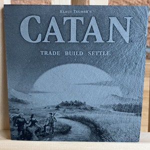 May include: A black and white image of a slate tile with the words "Klaus Teuber's Catan Trade Build Settle" printed on it. The image also features a stylised landscape with a large sun, a road, and three figures.