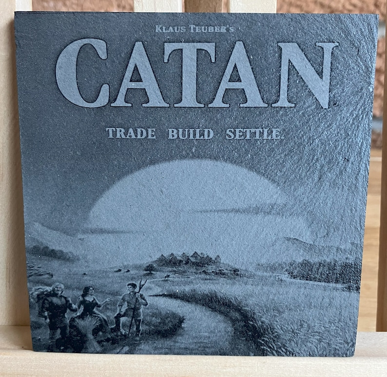 May include: A black and white image of a slate tile with the words "Klaus Teuber's Catan Trade Build Settle" printed on it. The image also features a stylised landscape with a large sun, a road, and three figures.
