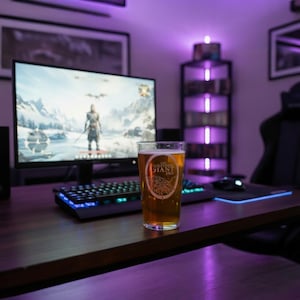 Op de afbeelding: Een pintglas gevuld met bier staat op een houten bureau voor een computermonitor. Het glas heeft een logo met de tekst "Sleeping Giant Inn". Een toetsenbord en muis staan ook op het bureau. De kamer is verlicht met paars licht.