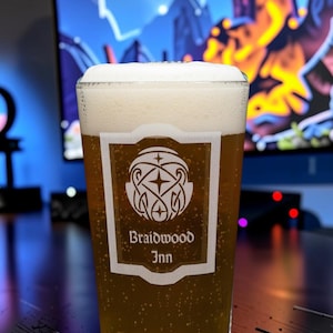 Peut inclure: Une pinte en verre transparent remplie de bière dorée surmontée d'une mousse blanche. Le verre présente un emblème rectangulaire blanc avec un motif de nœud en forme d'étoile et le texte Braidwood Inn imprimé en police sombre.
