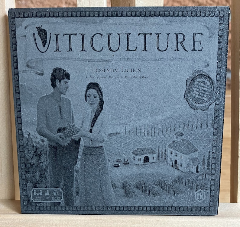 May include: A black and white image of a board game box with the title "VITICULTURE ESSENTIAL EDITION" printed on the front. The box features a couple standing in a vineyard with a small village in the background.