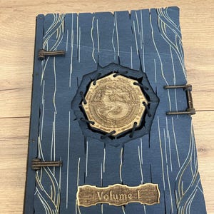 Peut inclure: Un journal en bois bleu foncé avec une couverture en bois sculptée décorative. La couverture présente un motif circulaire avec une bordure cousue et le texte "Volume I" sur une petite plaque en bois.