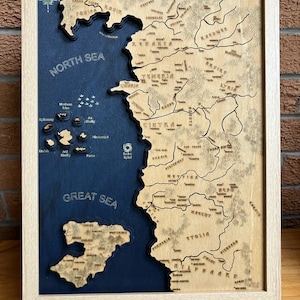 Könnte beinhalten: Eine Holzkarte von Westeros, einem fiktiven Kontinent aus der beliebten Fantasy-Serie Game of Thrones. Die Karte ist in einem hellen Holzrahmen gerahmt und hat einen blauen Hintergrund mit erhabenen Holzdetails. Die Karte enthält die Namen verschiedener Regionen und Orte, wie zum Beispiel die Nordsee, das Große Meer und die Eiseninseln.