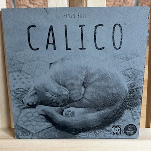 May include: A grey board game box with the title "Calico" printed in black. The box features a black and white illustration of two cats curled up together. The box also includes the text "Kevin Russ" and "Illustrated by Beth Sobel".