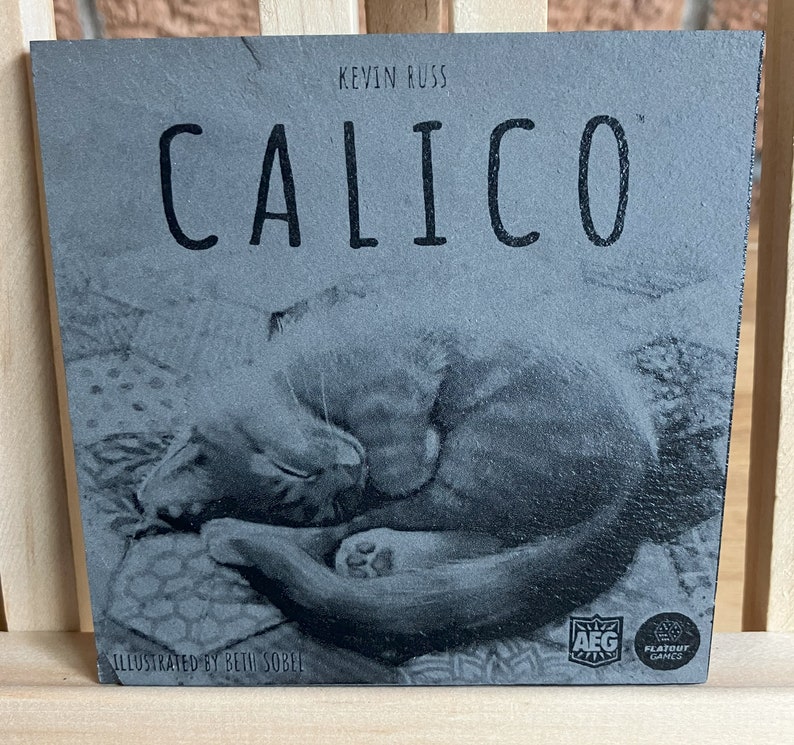 May include: A grey board game box with the title "Calico" printed in black. The box features a black and white illustration of two cats curled up together. The box also includes the text "Kevin Russ" and "Illustrated by Beth Sobel".