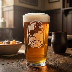 Op de afbeelding: Een helder glazen pint gevuld met goudkleurig bier en een witte schuimkraag. Het glas heeft een wit embleem met het silhouet van een steigerend paard en de tekst Prancing Pony in het zwart gedrukt. Het glas staat op een houten ondergrond.
