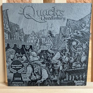 May include: A black and white illustration of a medieval town square with a sign that reads "The Quacks of Quedlinburg". The image is on a grey background with a North Star Games logo in the bottom right corner. The image also includes a 2018 Kennerspiel des Jahres award.