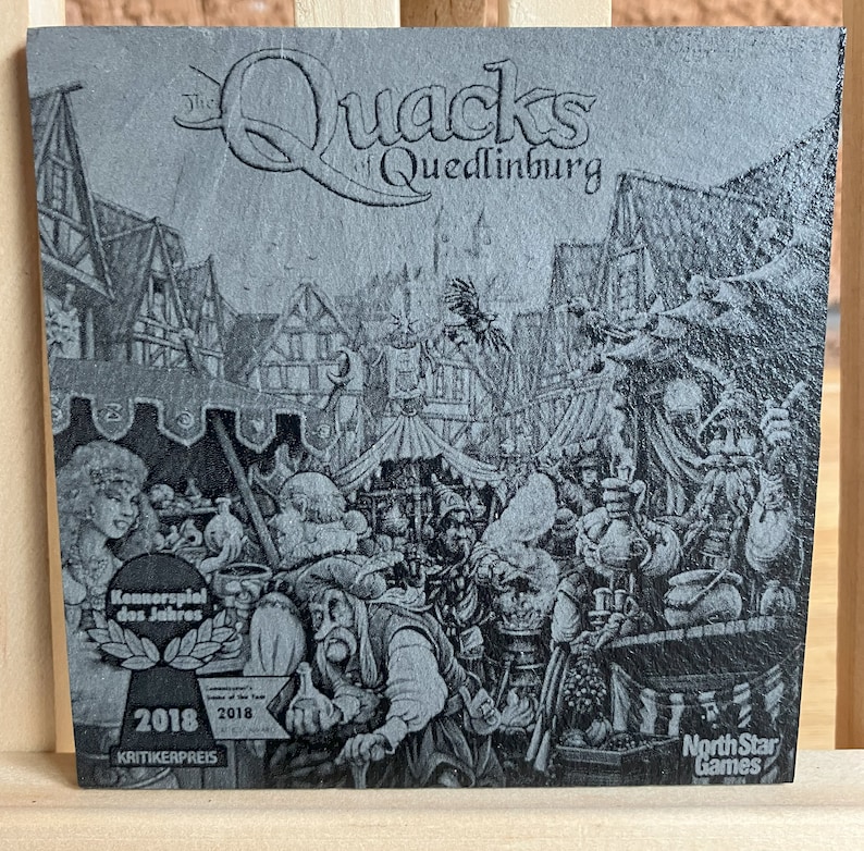 May include: A black and white illustration of a medieval town square with a sign that reads "The Quacks of Quedlinburg". The image is on a grey background with a North Star Games logo in the bottom right corner. The image also includes a 2018 Kennerspiel des Jahres award.