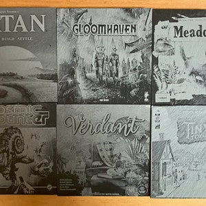 May include: A collection of nine grey slate coasters featuring various board game titles, including Catan, Gloomhaven, Meadow, Cosmic Encounter, Verdant, and Tiny Towns.