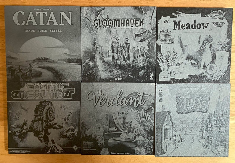 May include: A collection of nine grey slate coasters featuring various board game titles, including Catan, Gloomhaven, Meadow, Cosmic Encounter, Verdant, and Tiny Towns.