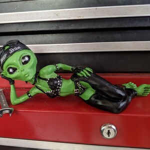 Op de afbeelding: Groene alien-figuur met een zwart leren outfit en een bandana met de tekst "Outer Babe". De figuur ligt op een rood oppervlak.