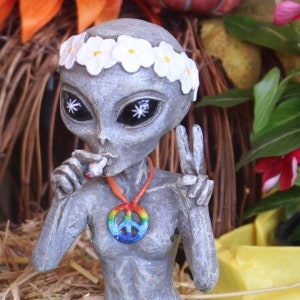 K&ouml;nnte beinhalten: Eine graue Alien-Figur mit einer Friedenszeichen-Halskette und einer Blumenkrone. Das Alien h&auml;lt eine Zigarette in der einen Hand und macht mit der anderen ein Friedenszeichen.