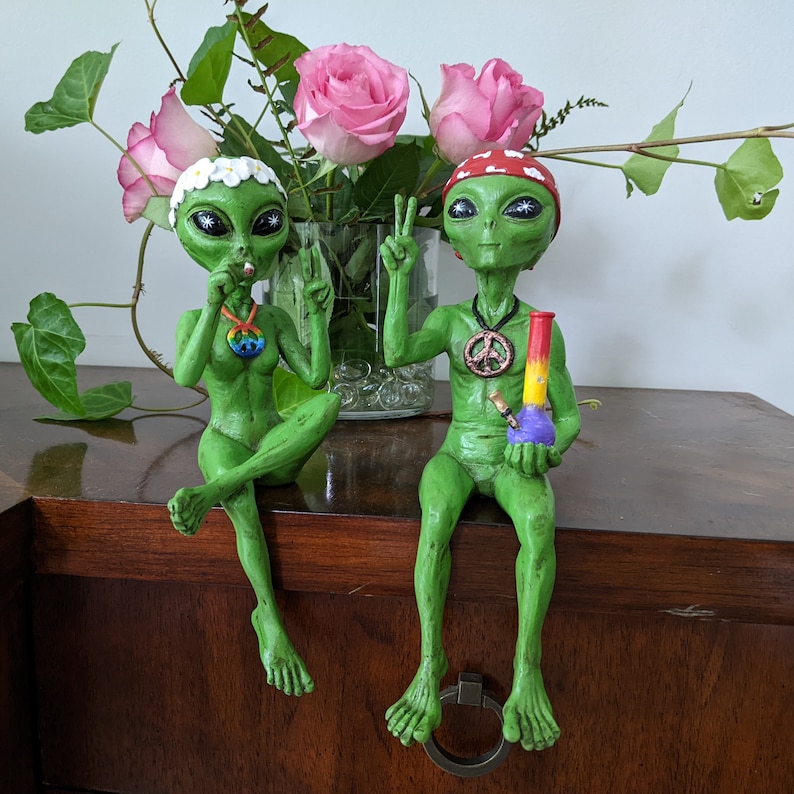 K&ouml;nnte beinhalten: Zwei gr&uuml;ne Alien-Figuren mit Friedenssymbolen um den Hals. Ein Alien h&auml;lt einen Joint und das andere eine Bong. Beide Aliens tragen Blumenkr&auml;nze.