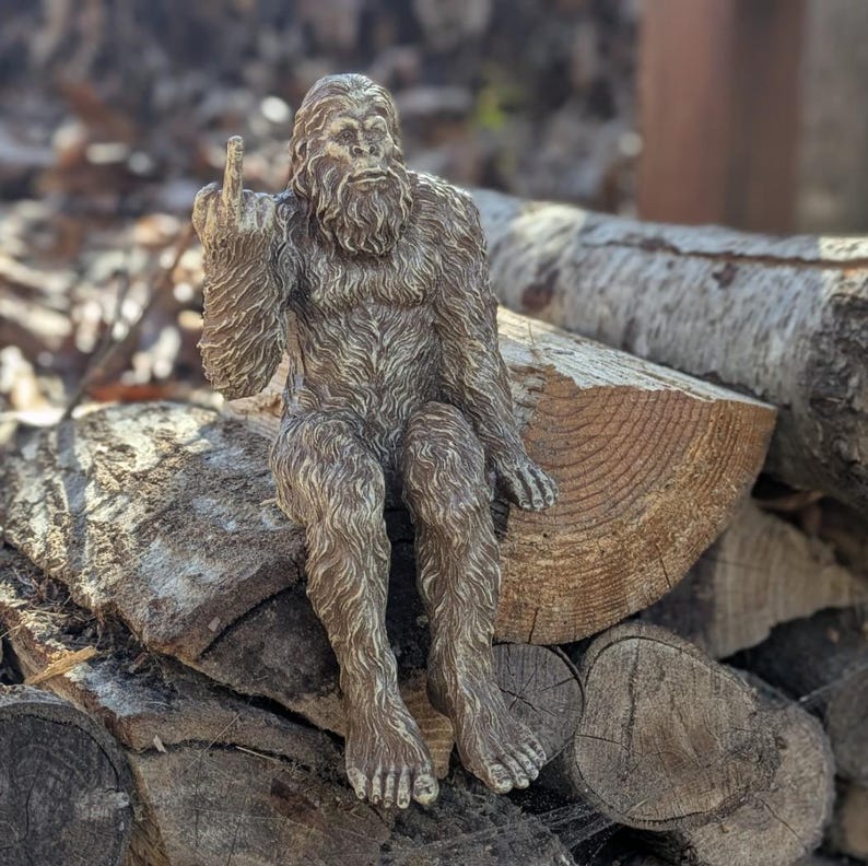May include: A brown, furry Bigfoot figurine sitting on a pile of logs. The figurine is giving the middle finger.