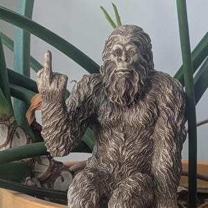May include: A brown and gray figurine of a Bigfoot giving the middle finger. The figurine is sitting on a pot of green plants.