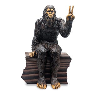 May include: A dark brown, hairy Bigfoot figurine sitting on a textured, dark brown log. The creature has a light-colored face, hands, and feet, and is holding its left hand up in a peace sign gesture with two fingers extended upward.