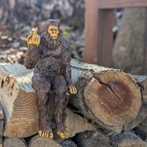 May include: A brown, furry figurine of a Bigfoot sitting on a pile of logs. The figurine is giving the middle finger.