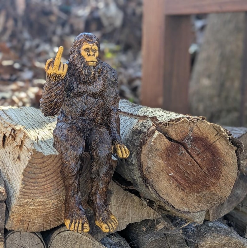 May include: A brown, furry figurine of a Bigfoot sitting on a pile of logs. The figurine is giving the middle finger.