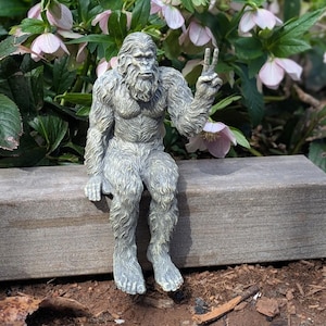May include: A gray, textured figurine of a hairy, ape-like creature sitting with its legs dangling. The creature has a thick coat of fur, a prominent brow, and is holding one hand up with two fingers extended in a peace sign gesture.