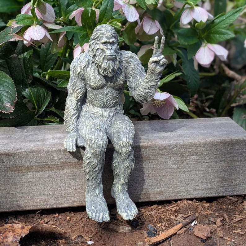 May include: A gray, textured figurine of a hairy, ape-like creature sitting with its legs dangling. The creature has a thick coat of fur, a prominent brow, and is holding one hand up with two fingers extended in a peace sign gesture.