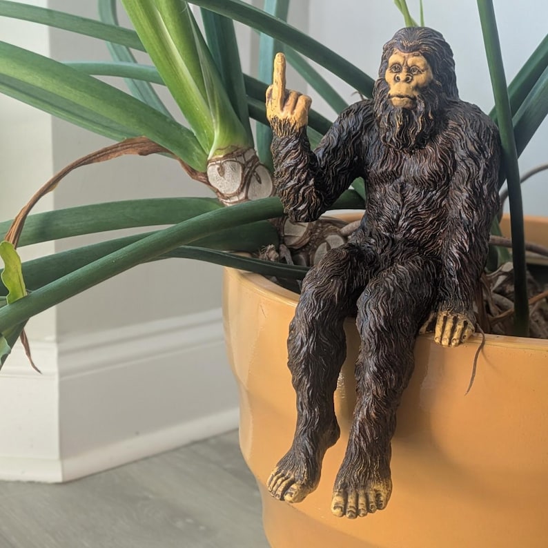 May include: A brown and black Bigfoot figurine sitting on a pot with a green plant. The figurine is giving the middle finger.