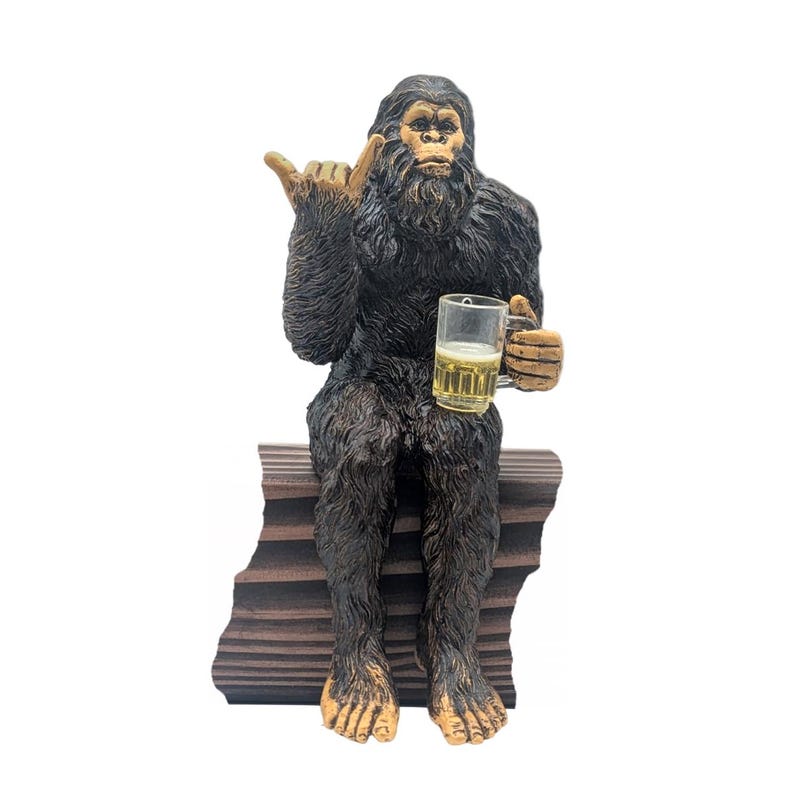 May include: A dark brown Bigfoot figurine with a beer mug, giving a 'hang loose' hand gesture. The figurine is seated on a wooden bench. The beer mug is filled with a light yellow liquid.