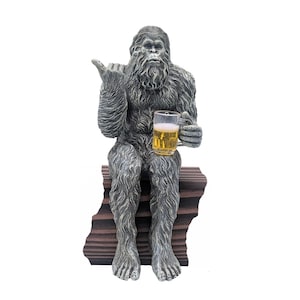 May include: A gray Bigfoot statue with a beer mug, seated on a wooden base. The statue has a furry appearance and is giving a thumbs-up gesture. The beer mug is filled with a golden liquid.
