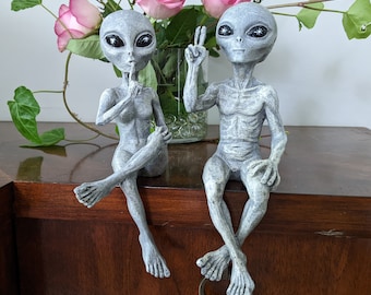 May include: Two gray alien figurines, one with a finger to its lips and the other with a peace sign, sitting on a wooden surface.