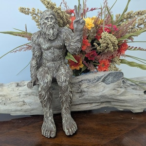May include: A gray, textured figurine of a hairy, bipedal creature with a beard, sitting down and holding one hand up in a peace sign gesture. The creature has detailed fur texture and is positioned in front of a colorful floral arrangement.