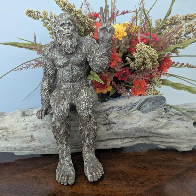May include: A gray, textured figurine of a hairy, bipedal creature with a beard, sitting down and holding one hand up in a peace sign gesture. The creature has detailed fur texture and is positioned in front of a colorful floral arrangement.