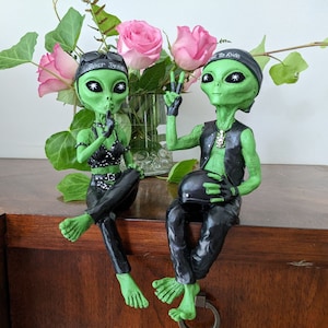 Op de afbeelding: Twee groene alien-figuurtjes die zwarte leren jacks en helmen dragen. Het figuurtje links houdt een vinger op zijn lippen en het figuurtje rechts houdt een helm vast. Beide figuurtjes hebben groene huid en zwarte ogen.