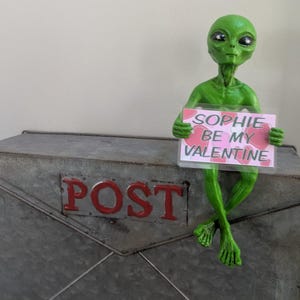 May include: A green alien figure holding a sign saying "SOPHIE BE MY VALENTINE." The alien is sitting on a metal postbox with the word "POST" in red. The background is a plain white wall.
