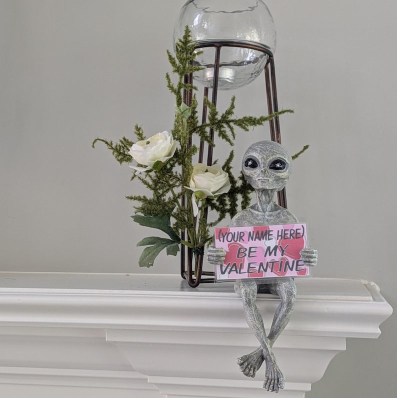 May include: A grey alien ornament holding a sign saying "(YOUR NAME HERE) BE MY VALENTINE." The alien sits on a white surface, with a decorative metal stand holding a glass globe and greenery with white flowers in the background.