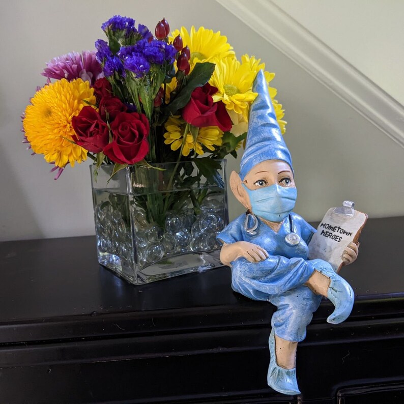 Op de afbeelding: Een blauwe dwergfiguur met een chirurgisch masker en een schort, die een klembord vasthoudt met de tekst "Hometown Heroes". De dwerg zit op een tafel met een vaas met kleurrijke bloemen.