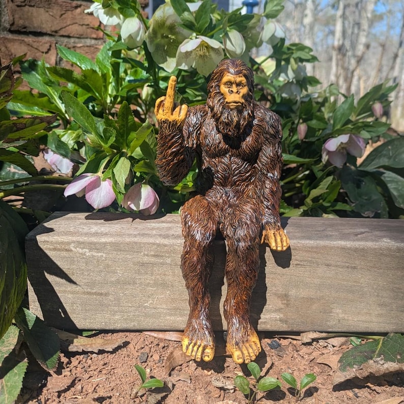 May include: A brown, furry Bigfoot figurine sitting on a wooden ledge. The figurine is giving the middle finger with its right hand.